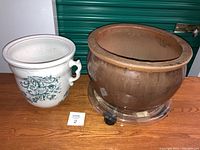 Photo showing both planters side by side on wooden surface; left is white porcelain with green floral design, right is larger brown earthenware with rolling tray below.