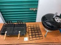 Two small tabletop grills on a wooden table: one Weber round black grill and one rectangular grill with wooden handles, accompanied by two extra cooking grates.