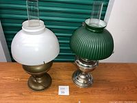 Two hurricane lamps on wooden table against green storage unit background. Left lamp with white opaque globe shade, brass-colored base. Right lamp with ribbed green globe shade, silver-colored base. Both have clear glass chimneys.