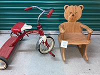 Photo showing the Radio Flyer tricycle next to the wood teddy bear rocking chair on concrete floor with a teal roller door in background.