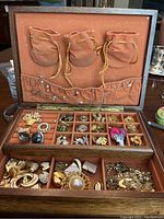 Open wooden jewelry box showing multiple compartments filled with assorted vintage costume jewelry including brooches, earrings, necklaces, chains, and pins.