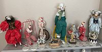 Full view of 9 clown collectible items displayed on a shelf including dolls on stilts, on tricycles, and small figurines