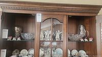 Wooden display cabinet with multiple shelves showing Dresden floral figurines, Dresden angels, cut glass bowl, matching cut glass pitcher and vase, and other porcelain and china plates.
