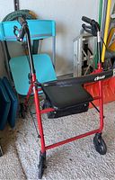 Red Drive brand folding walker with black padded seat, zippered storage bag underneath, and four wheels with black rubber tires. Part of a blue folding chair visible behind the walker.