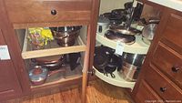 Open view of corner cabinet showing metal pots, pans, cast iron skillet, and mixing bowls stored inside wooden cabinetry