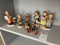 Shelf display of 5 Hummel figurines and 2 antique German dolls in assorted poses and outfits.
