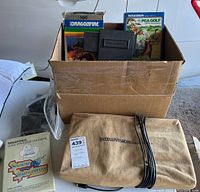 Photo showing a cardboard box containing game cartridges including Dragonfire and PGA Golf, and the Intellivision system inside a tan canvas carrying case with cables wrapped around it.
