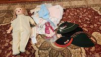 Overview of entire lot showing vintage baby doll, assorted antique crocheted baby clothes in pastel colors, and four traditional style felt hats in green and black with tassels and embroidery.