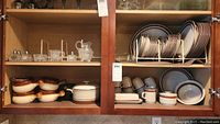 Shelves with various glassware on upper shelves and Dachi stoneware dish set on lower shelves, including plates, bowls, ramekins, and covered casserole dish