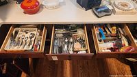 Three opened drawers displaying contents of mixed flatware and kitchen utensils, with visible Rebacraft bamboo pattern silverware and various miscellaneous kitchen tools.