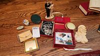 Photo showing various vintage compacts, small figurines, a porcelain figurine couple, and a bust figurine arranged on wood floor