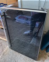 Front view of black wine fridge with glass door and silver handle, showing reflection of photographer.