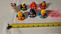 Photo showing all seven rubber ducks arranged in two rows with a ruler below for size reference, showing they measure about 3 inches long. Ducks are dressed in various superhero costumes, colorful and plastic/rubber material.