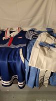 Stranger Things Robin costume laid out showing the blue dress with white sailor collar, red necktie, and white trim near the hem, next to the Dorothy costume's blue dress and white apron.