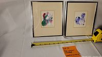Two framed original watercolor paintings side by side with tape measure showing size around 8 1/4" x 7 1/4"