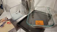 Photo showing clear plastic bread keeper, two metal graters, Pyrex glass baking dish, and small dark metal sheet pan stacked together with a blue pastry circle cutting set.