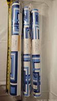 Three rolled peel and stick wallpaper pieces with a blue geometric pattern, measured next to a tape measure showing size.