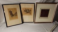 Three framed prints under soft light; two architectural images by Marjorie Christine Bates on cream paper and one smaller red abstract print.