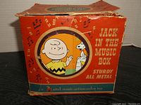 Replacement box for the 1953 Snoopy Jack in the Box toy, orange with music note graphics and Peanuts characters including Charlie Brown and Snoopy.