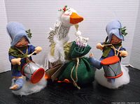 Three Annalee Dolls displayed: two blue-hatted Drummer Boys on drums and a white Golden Goose with a green cloth sack and orange beak.