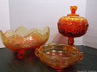 Three cranberry/amber glass dishes shown: a covered dish with scalloped lid and handle, a footed dish with scalloped edges, and a handled dish with embossed patterns.