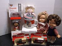 Full lot arrangement with dolls (boxed and loose), two Campbell's toy trucks in original packaging, and a Campbell's mug displayed on a white pedestal.