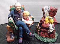 Figurines showing elderly man reading to a child in a green armchair and a red armchair with a sleeping dog, from a front angle.