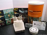 Photo showing the GAF Talking View-Master Gift Pak II storage container, boxes of talking reels 'Prehistoric Life' and another set, the power winder, reel cartridge, and stereo viewer.