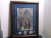 Framed and matted black and white 9/11 heroes print showing firefighters raising American flag among rubble with three commemorative stamps below