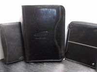 Photo of three black leather items: portfolio, day runner wristlet, and iPad case, all with visible zipper closures and embossed logos.