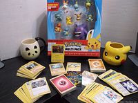 Overview photo of entire lot showing trading cards spread in piles, multi-pack figure set in box, and two character mugs.