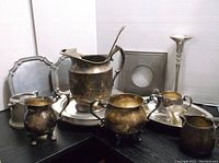 Photo showing multiple tarnished silver plate and pewter serving pieces including a large pitcher, creamer, sugar bowl, round tray, and a pedestal bowl.