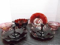 Full view of six cranberry glass pieces arranged showing handles, scalloped edges, and painted floral bowls.