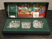 Three clear crystal train pieces in green velvet box with colorful holiday artwork inside lid and Christmas Train booklet, Marquis Waterford Crystal branding visible.