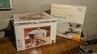 Photo showing boxed Seville Classics commercial chafing dish and Wolfgang Puck stainless steel mixing bowls on a wood surface against a basement wall.