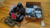 Overview of the lot including cameras, camera bags, and other accessories in a box and laid out on floor