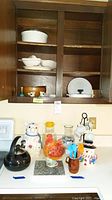 Several ceramic and glass kitchenware items are shown on a kitchen counter and wooden shelf including a Lenox pig cookie jar, Enesco chicken cookie jar, a snowman cookie jar, white casseroles, serving bowls, pitchers, salt and pepper shakers, and cups.