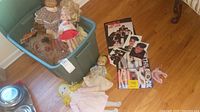 Image showing a green plastic bin containing several vintage dolls including a vinyl doll with blonde curly hair in a red and white outfit, a plush monkey, and other dolls. In front of the bin are New Kids on the Block souvenir books and photo cards.