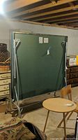 Folded green ping pong table standing upright in basement showing metal frame and legs with wear and rust.