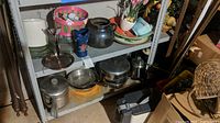 Multiple Revere Ware sauce pans, fry pans, stock pot, coffee pot, plastic fruit decor, basket, plates, and vases placed on metal shelves in basement with visible signs of dust and rodent evidence.