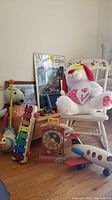 Photo showing a white wooden rocking chair with pink floral paint, a red and white wooden doll cradle, vintage Fisher Price Pull A Tune xylophone toy, music clock teaching toy, plush dog toy, wooden airplane toy, and other small toys and framed pictures in the background.