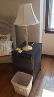 Gold tone table lamp atop black metal mesh rolling cart with white wicker trash basket nearby on wooden floor.