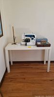 Full view of the sewing machine placed on the white Parsons style table alongside sewing boxes and a height gauge tool.