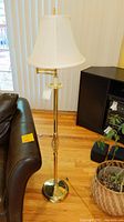 Full view of a brass-toned metal swing arm floor lamp with white fabric shade, standing on wooden floor beside dark furniture and a couch.