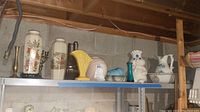 Top shelving view showing an array of vases, cookie jar, ceramic bowl and pitcher in various colors and styles on a white shelf against concrete basement wall.