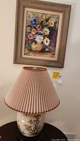 View of entire lamp with pleated shade and floral painting in wood frame hung on wall above it.