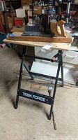 Photo of black metal Task Force foldable work table with wooden top and Stanley wooden miter box mounted on it, taken in basement workshop environment.