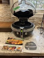 Front view of the T-Fal Actifry 2 in 1 appliance showing open lid, internal stirring paddle, and included cookbook/manual on countertop.