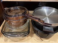 Multiple pieces of clear glass bakeware stacked: saucepan with handle and lid; casserole dishes; and a large metal roasting pan alongside.