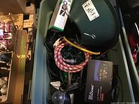Lidded green plastic bin containing various Christmas electrical items: green LED lights, rope lights coiled in red and white patterns, tree stand and laser projector box.
